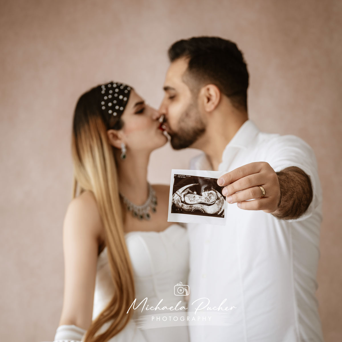 Paar im Studio küsst sich liebevoll, während der Mann ein Ultraschallbild ihres Babys in die Kamera hält.